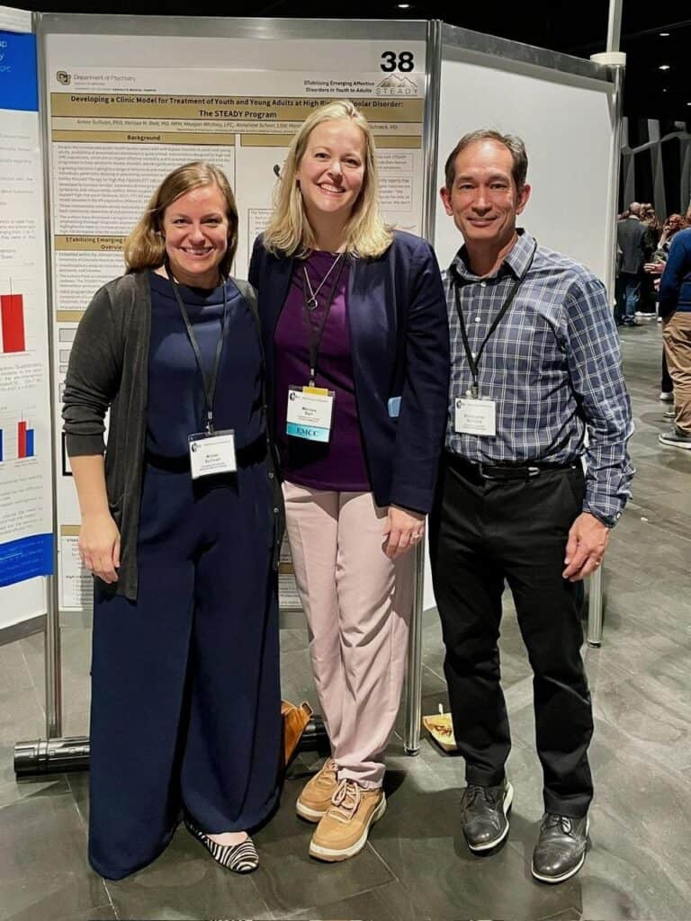 Three people stand smiling in front of a research poster at a conference.